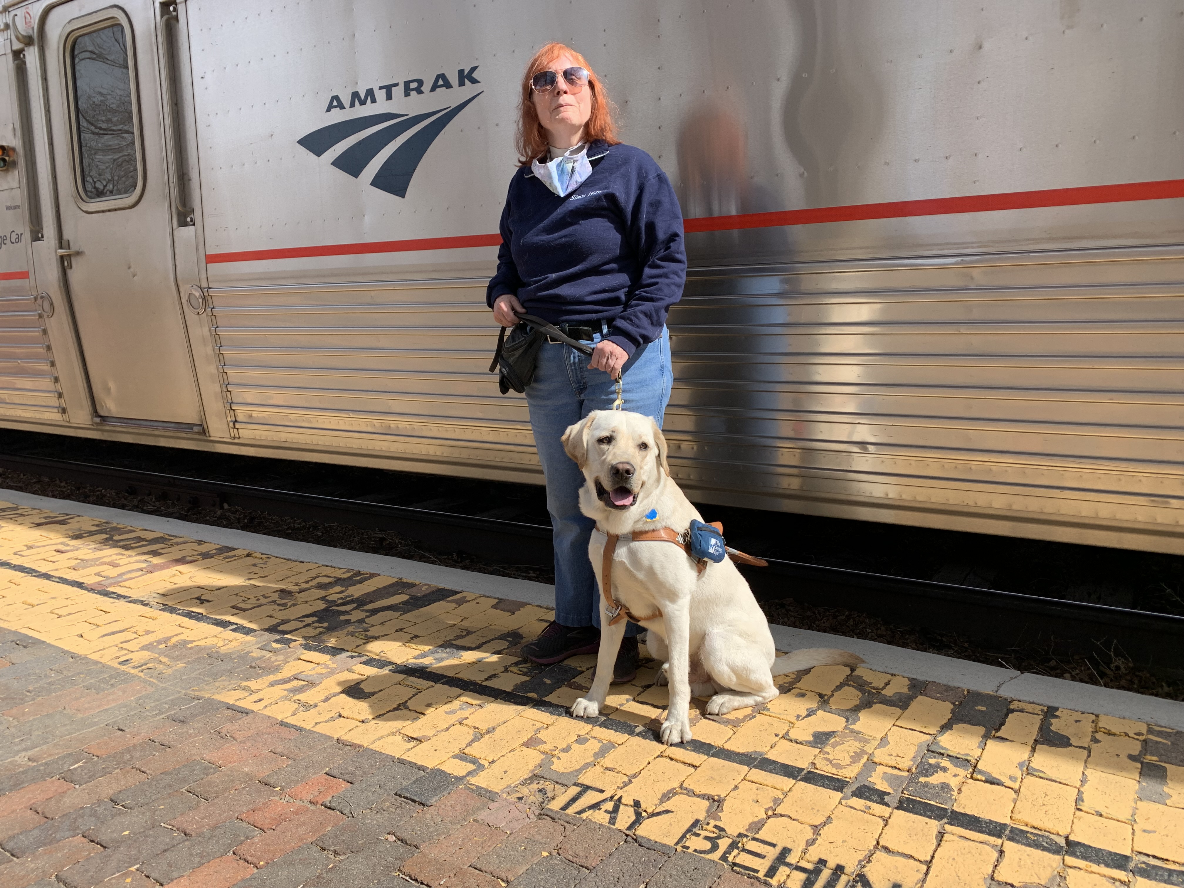 Karen stands with her new Seeing Eye dog on a platform in front of a gleaming Amtrak coach