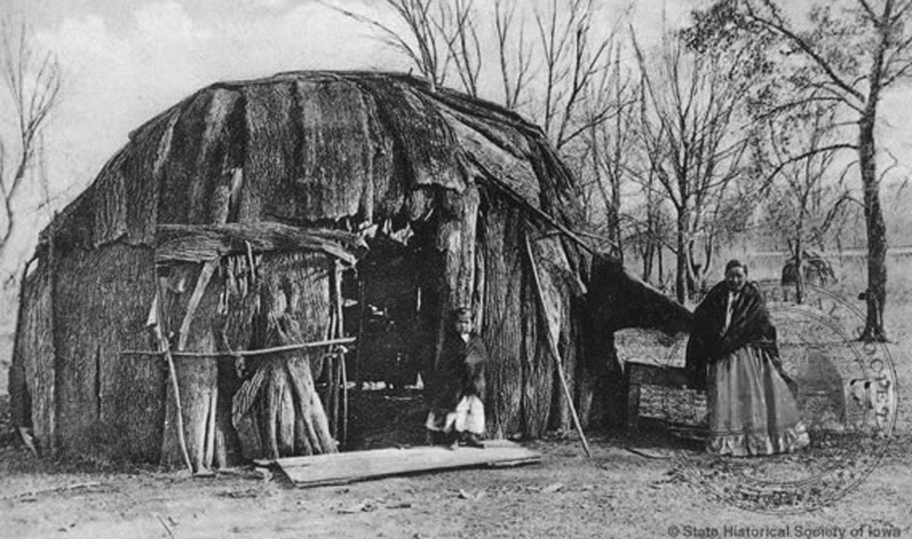 : A woman and girl stand in front of a Meskwaki wickiup. These dome-shaped structures, covered with slabs of tree bark, were winter quarters for family groups.