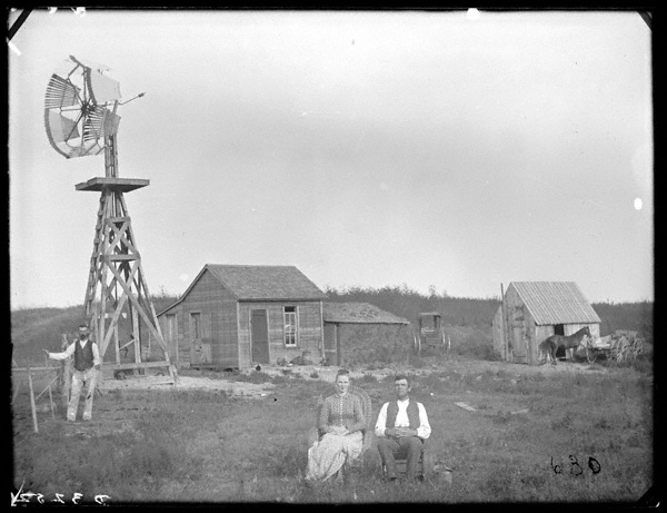 Old black and white photo of a prairie farmstead. Small, rude frame house and large wooden windmill and small livestock building and wagon in the background. Man and woman seated in the foreground and another man stands with his hand on a fence near the windmill.