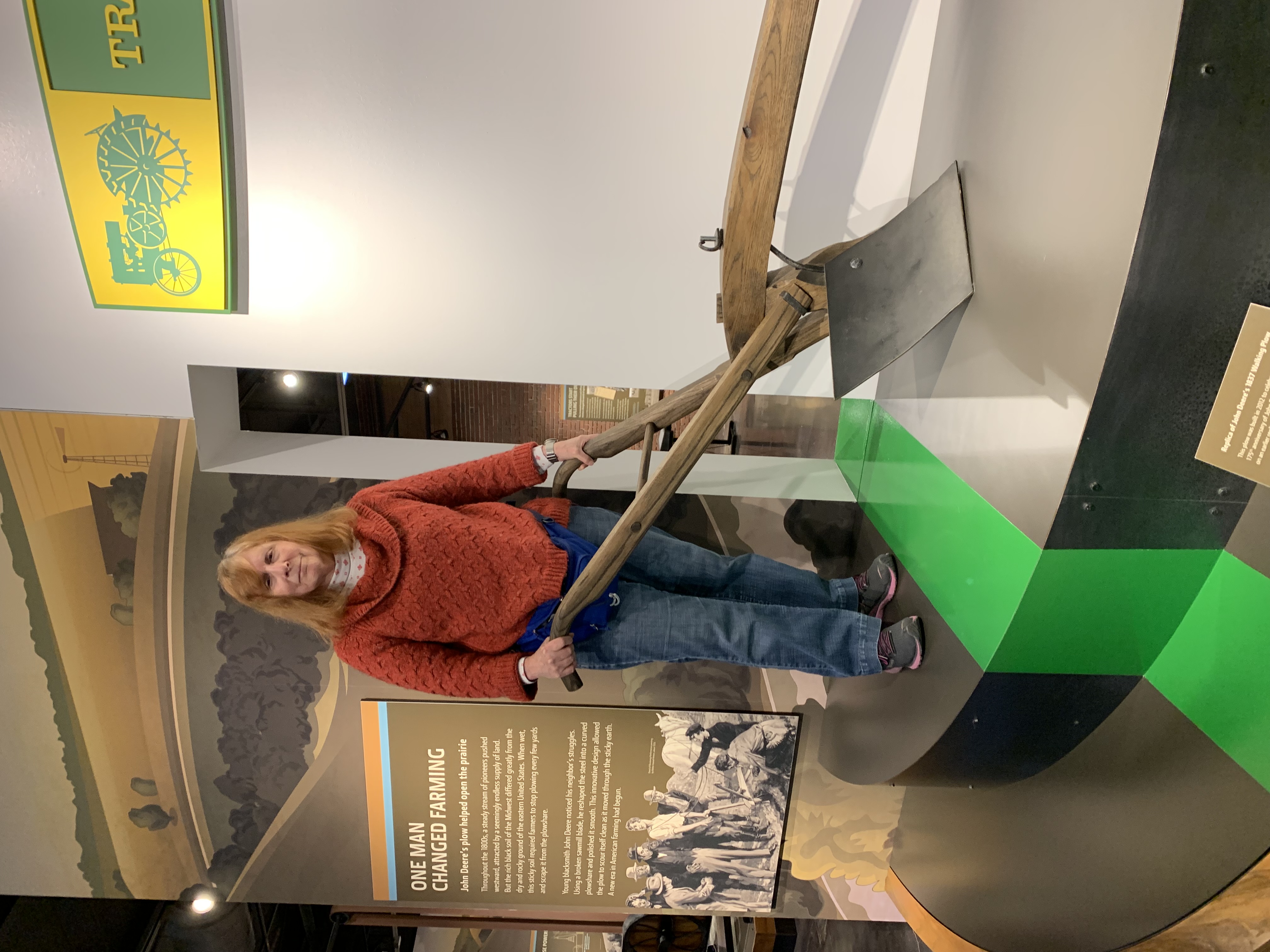 Karen stands behind a replica of John Deere’s sod-busting plow he invented in 1837. A stout wooden beam supports a curved, diamond-shaped plowshare, made from a sawmill blade. The point cuts the sod and starts it curving up and across the face of the plowshare. As the sod slides across the steel, it continually scours the surface clean so the plow doesn’t get clogged by the sticky prairie soil.