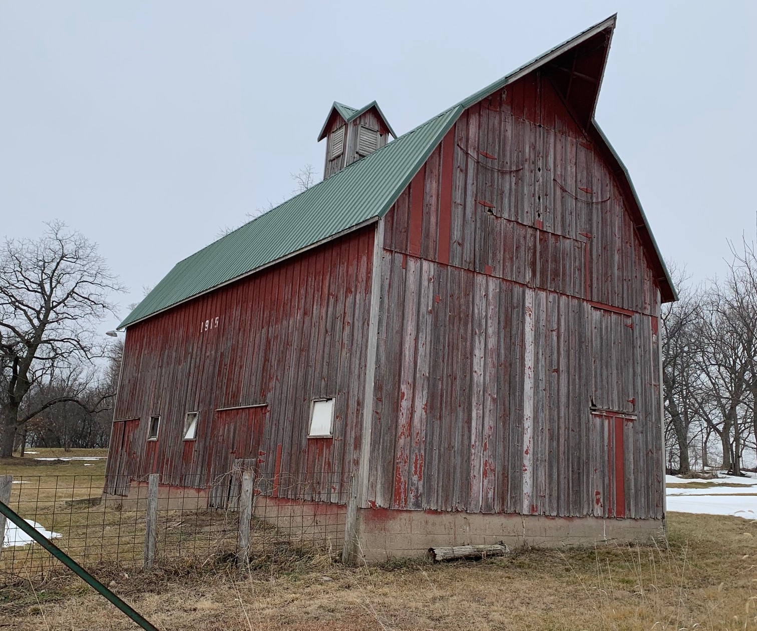 The 1915 red barn on Owl Acres. Weathered, board-and-batten siding and green metal roof. Four-sided cupola with louvered panels perches on top. The roof has a sharp, pointed extension over the east gable, housing the track for the hay conveyor and providing partial cover for the large hay doors.
