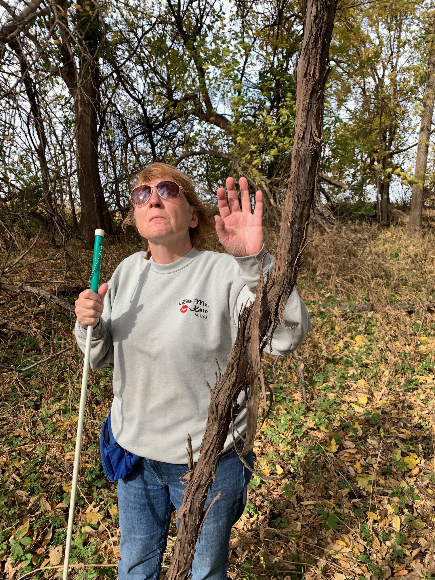 Karen is in the woods behind the house on a bright fall day. The trees have dropped most of their leaves. A large woody vine rises from bottom to top clear through the frame. It's the main stem of a river grape, its branches and foliage tangled high overhead in the nearby tree.